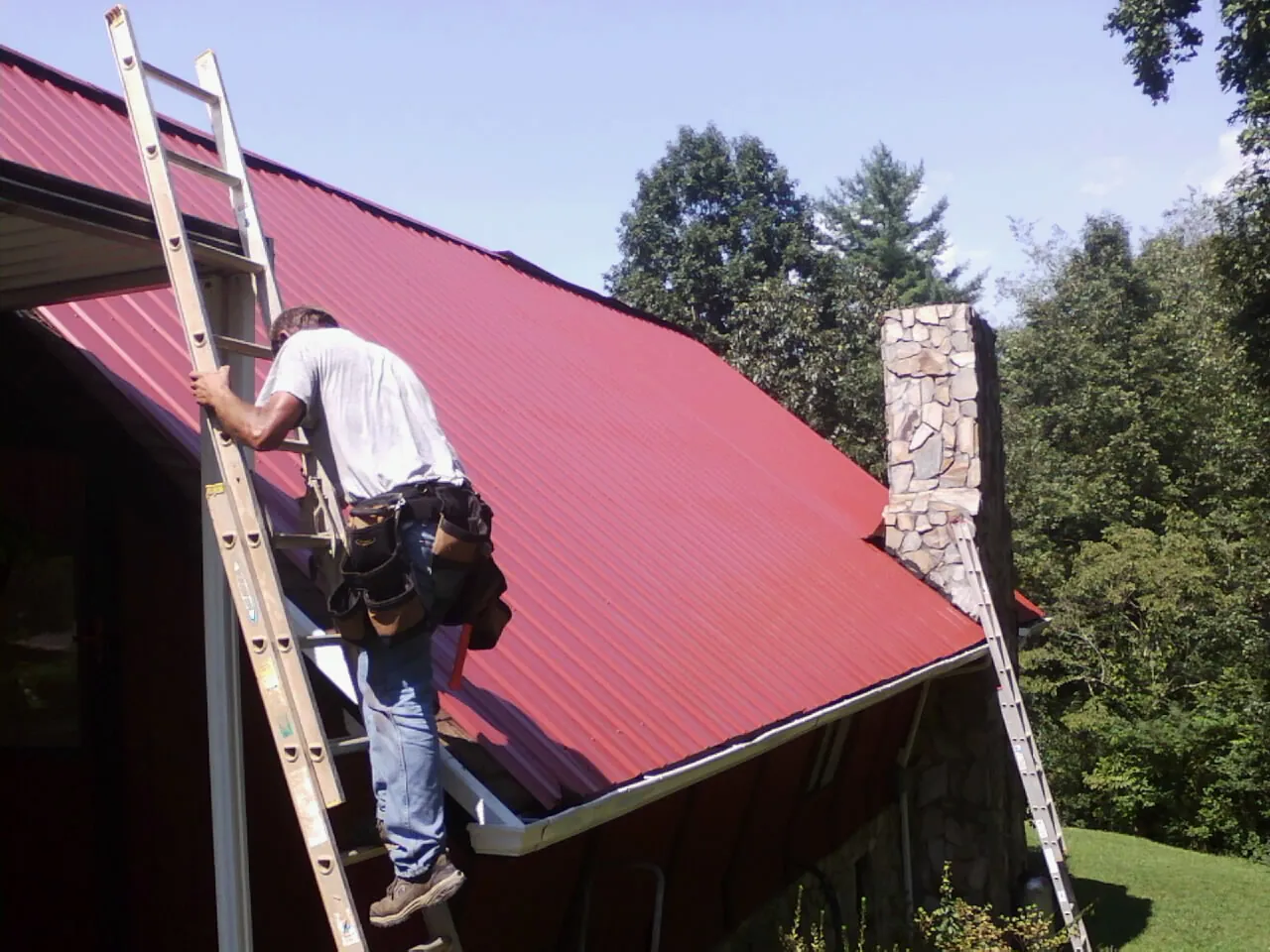 Roofer on a ladder beside a red metal roof and stone chimney — Soffit & Fascia Repair by Roofing Brothers in Santa Clarita